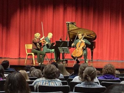 A musical trio on a stage with a red curtain backdrop in front of a seated crowd.