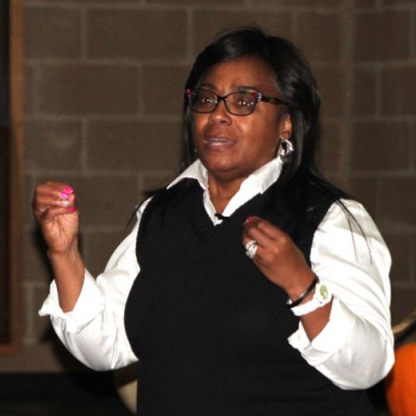 A woman with dark hair talking in a white shirt and black sweater vest with glasses.
